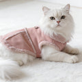 White fluffy cat wearing a pink fleece pet jacket on a soft white carpet